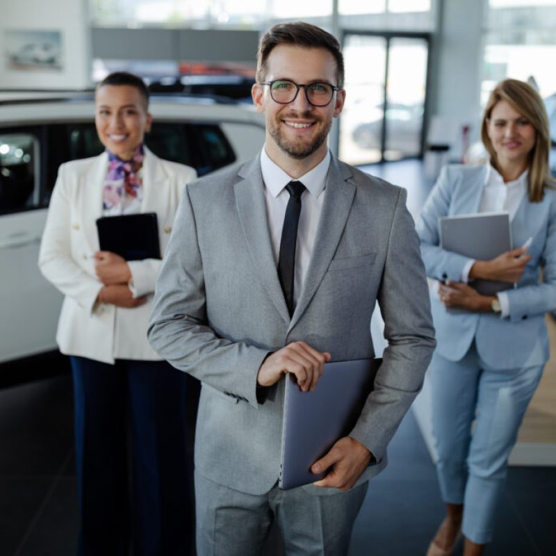 Confident salesteam in dealership, three beautiful consultants or managers in elegant suit looking on camera.